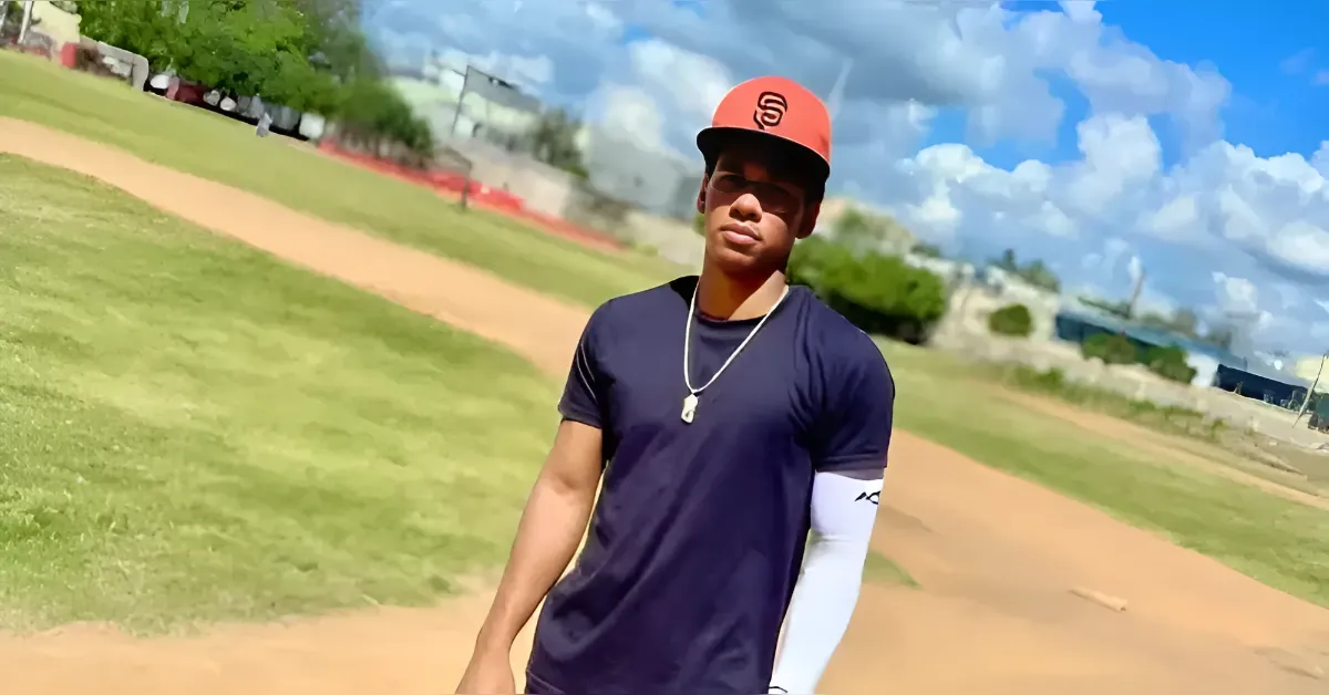 Young man in an orange cap and dark T-shirt stands on a dusty baseball field, facing the camera.