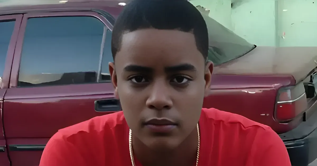 Close-up of a young man with a short haircut wearing a red shirt, standing in front of a maroon car.