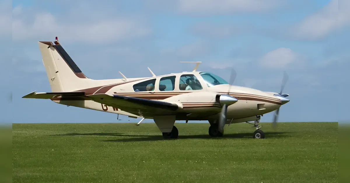 Small twin-engine propeller aircraft parked on a grassy airfield under a blue sky with a light cloud cover to the left, nose facing right.