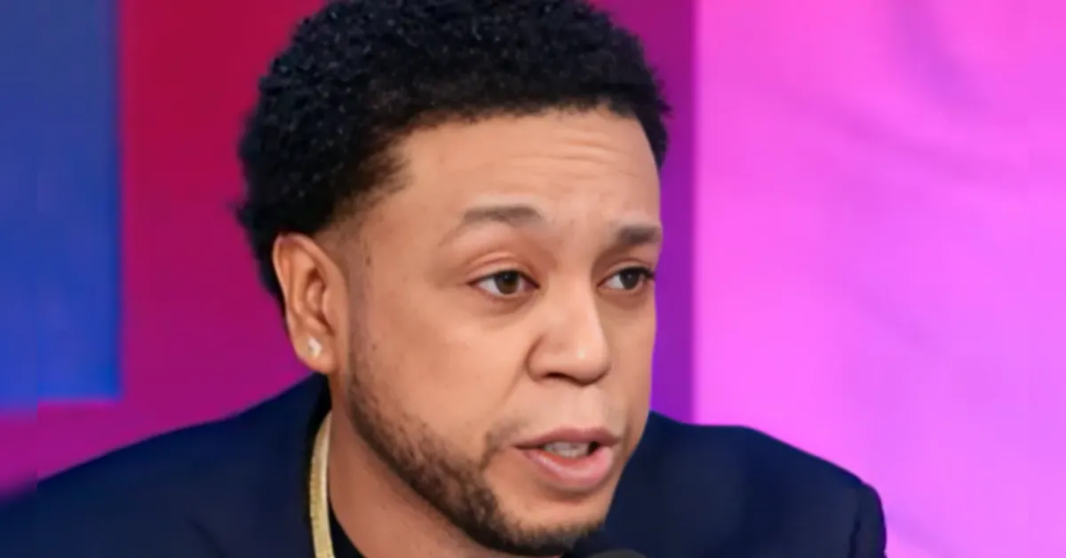 Close-up of a man with short black hair and an earring speaking into a microphone against a pink and purple background.