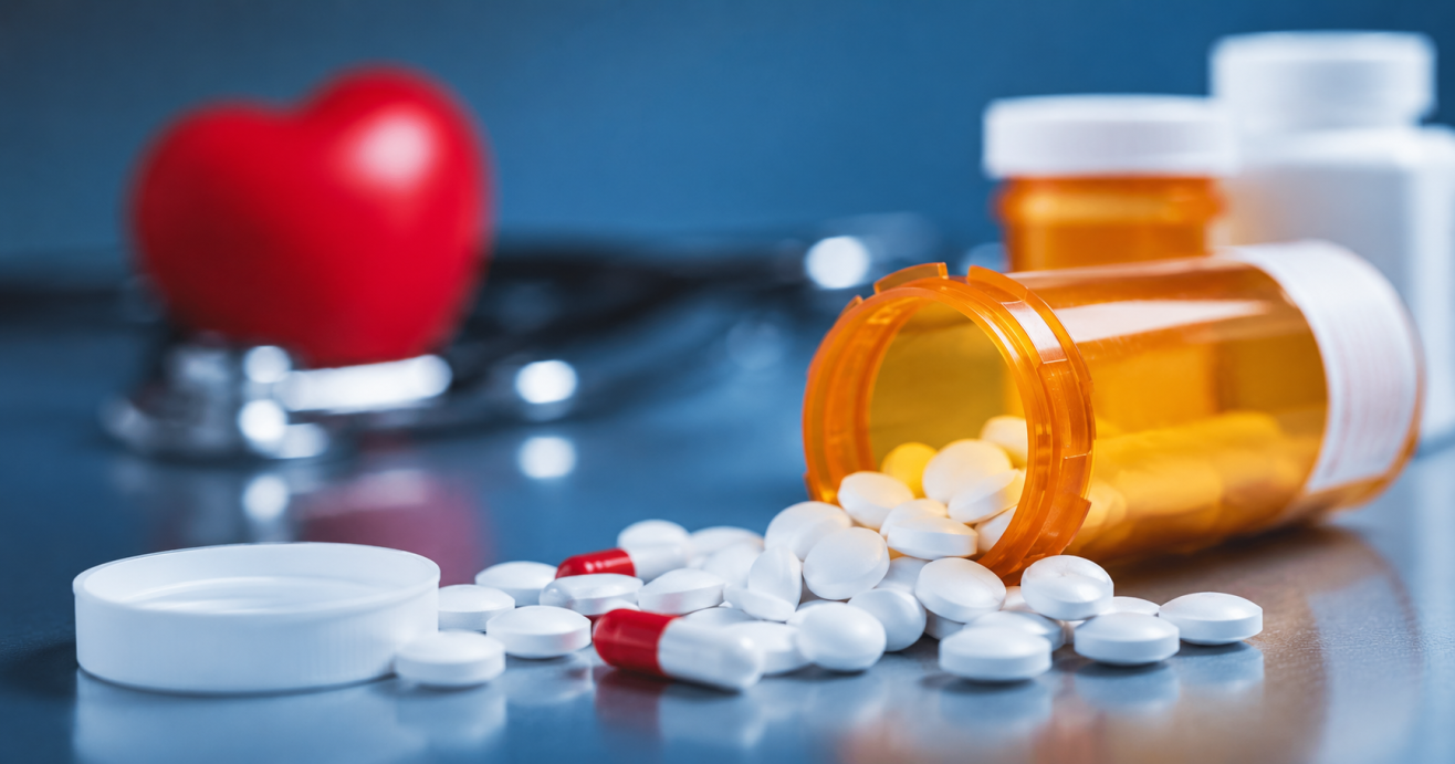 Prescription bottle spills white tablets and red capsules on a clinic counter, with a stethoscope and a red heart blurred in the background; medicine and heart health theme.