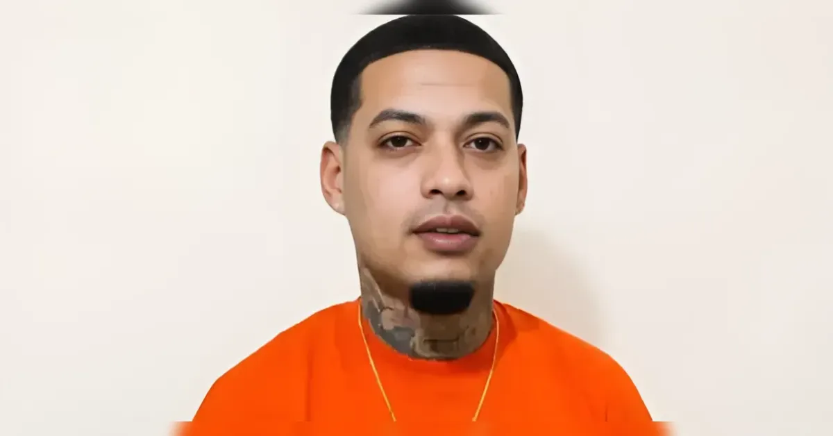 Head-and-shoulders image of a man in an orange shirt with visible neck tattoos, standing against a light background.