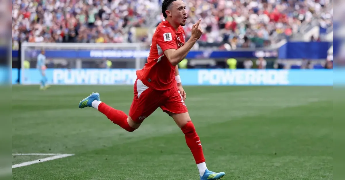 Soccer player in a red kit celebrating a goal, running on the field with a finger raised toward his lips as fans cheer in the background