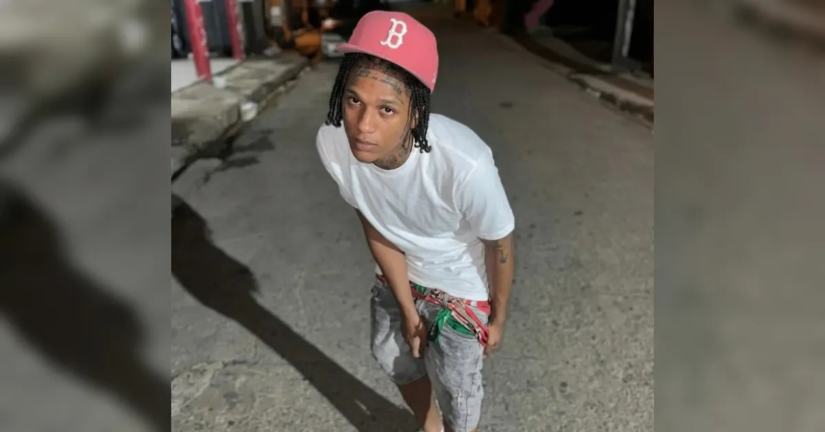 Young man in a white T-shirt and gray shorts with a red baseball cap, standing in an urban alley and looking at the camera.