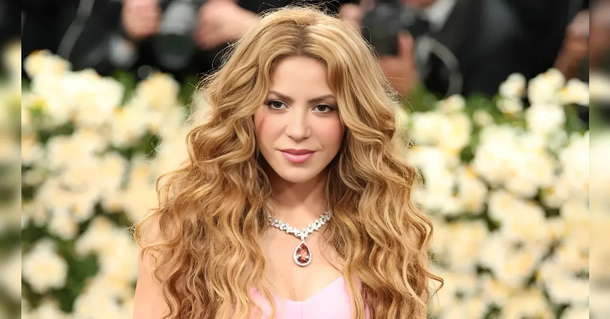 Woman with long, wavy blonde hair wearing a pink dress and a diamond teardrop necklace at a floral backdrop, posing for the camera on a red carpet-style event.