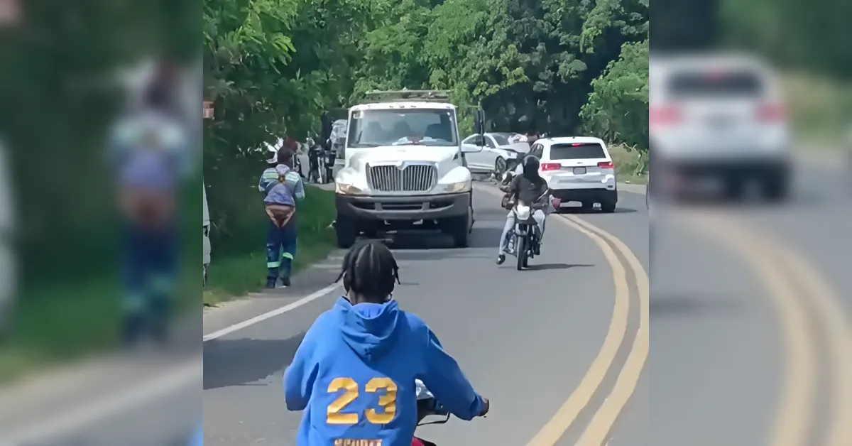 A white large truck blocks a two-lane rural road as cars and motorcycles follow; a rider in a blue hoodie with the number 23 is in the foreground.