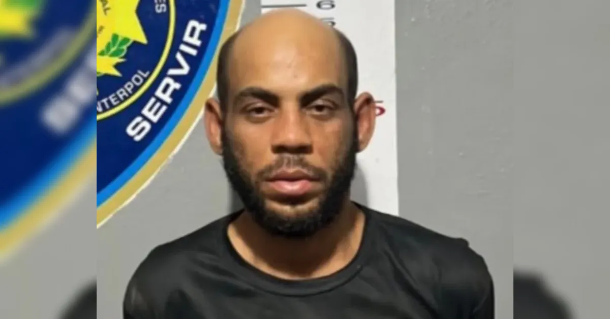 Mugshot of a man with a bald head and beard standing against a police backdrop with a badge partially visible behind him.