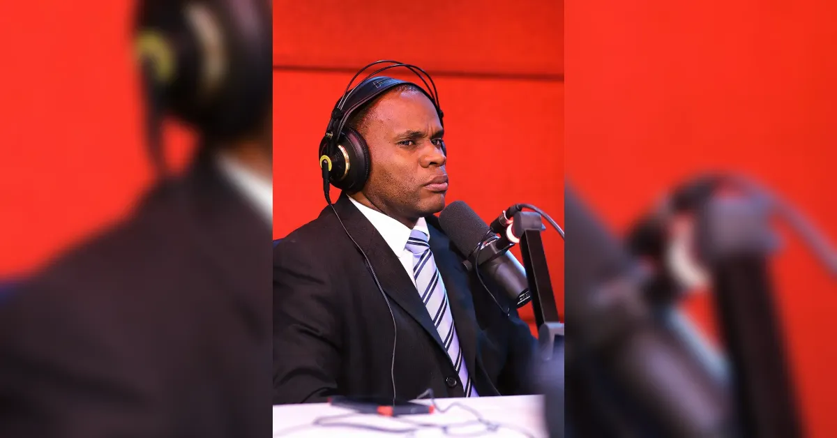Man in a suit wearing headphones speaks into a microphone in a recording studio with a bold red background backstage setup.