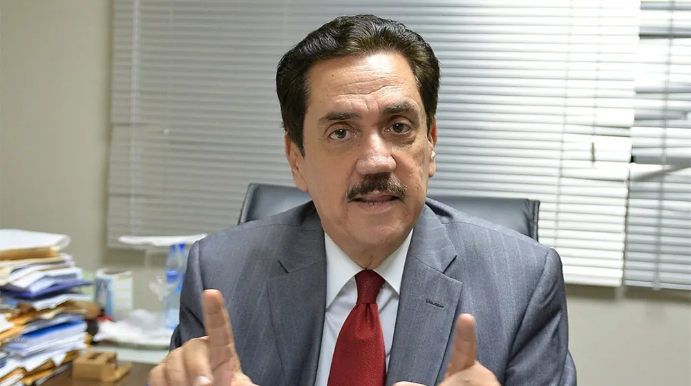 Man in a gray suit and red tie seated at a desk, gesturing with two raised fingers as he speaks.