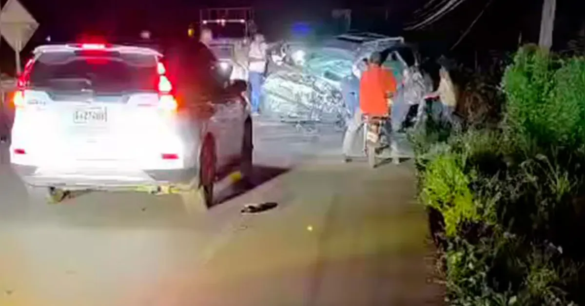 Nighttime accident scene with a white SUV stopped on a road, red and blue emergency lights flashing, and a group of people near a severely damaged vehicle ahead.