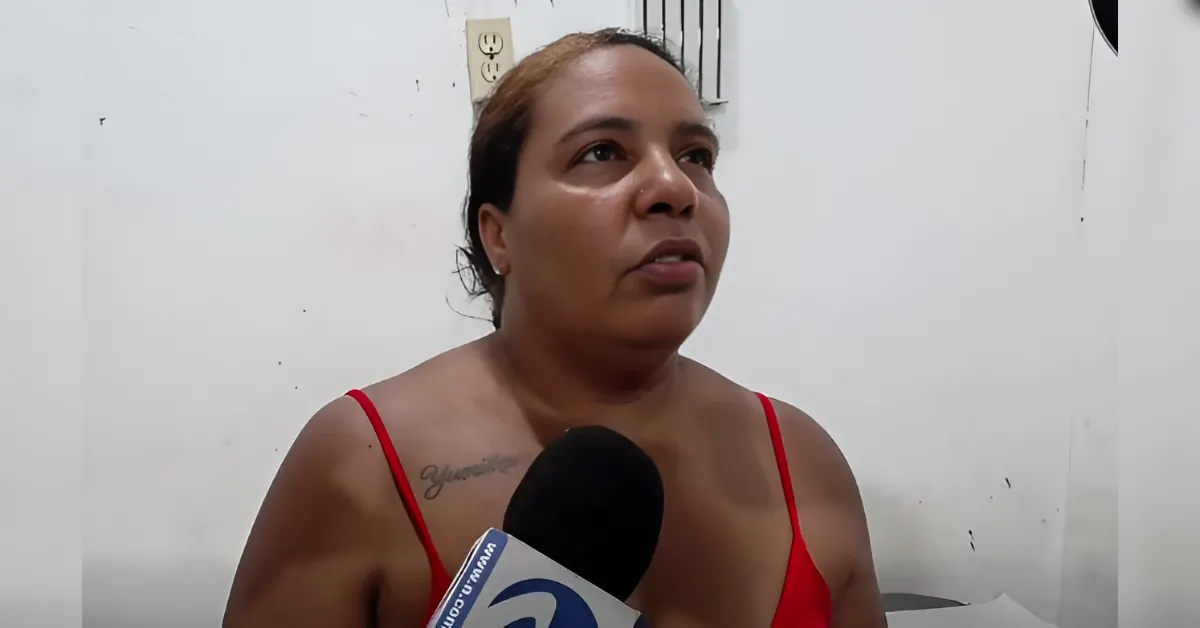 Woman in a red sleeveless top speaks into a handheld microphone during an interview, standing in a plain room with a white wall behind her.