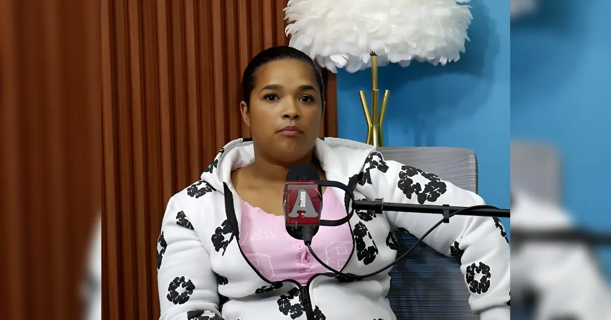 Woman seated at a podcast microphone, wearing a pink shirt and white hoodie with black floral patterns, against brown curtains and a blue wall.
