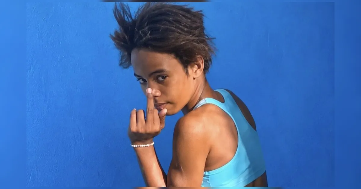 Person with short hair wearing a blue athletic top against a blue wall, flashing the middle finger at the camera.
