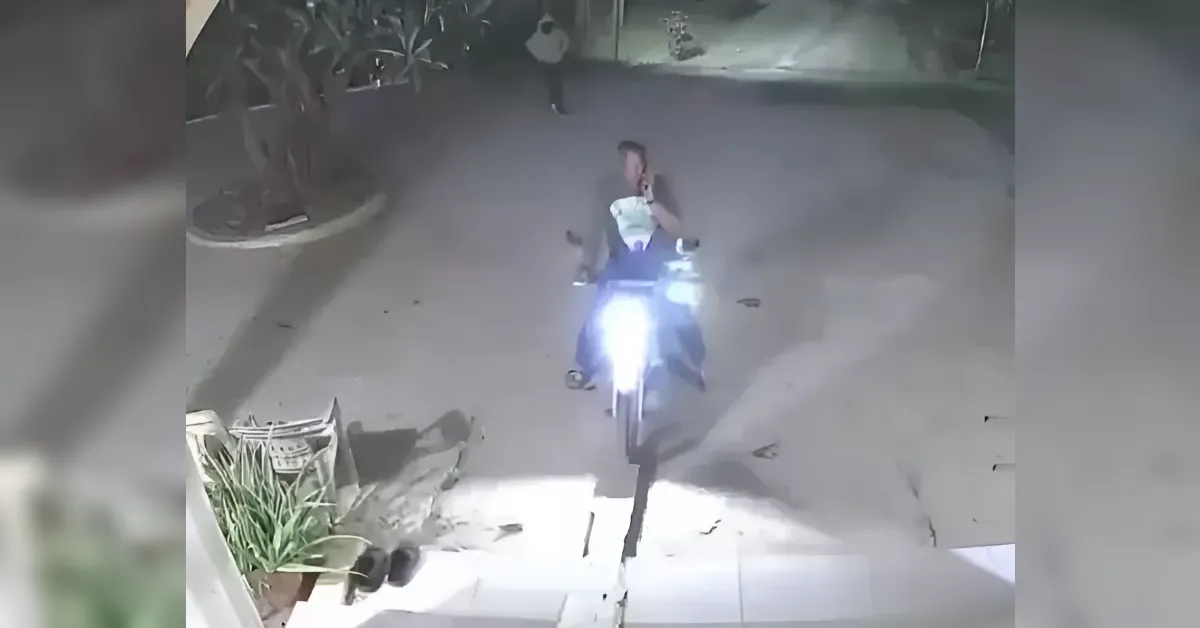 Motorcycle rider approaches the camera on a residential driveway at night; bright headlight and front steps with potted plants visible.