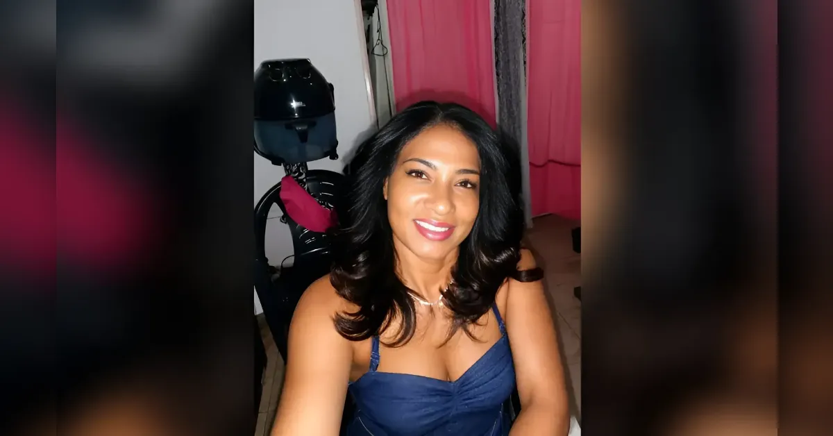 Smiling woman with dark hair in a blue dress taking a selfie indoors, pink curtains in the background.