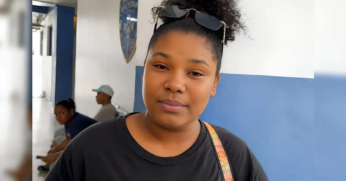 Portrait of a young woman with sunglasses on her head standing in a blue-and-white hallway with other people seated in the background.