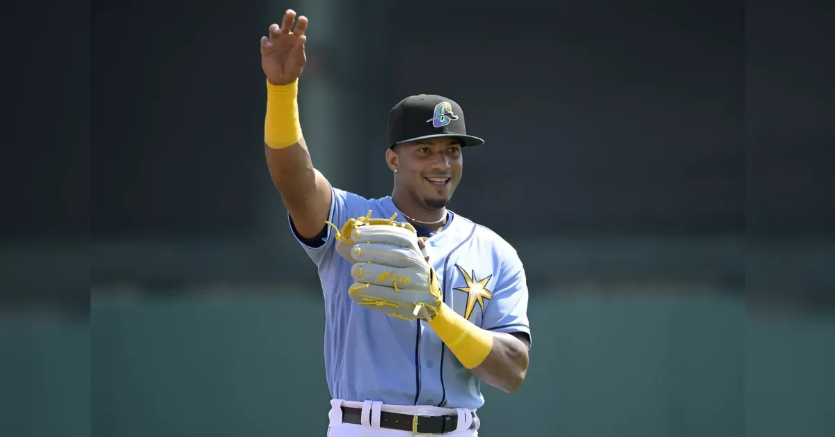 Baseball player in a light blue uniform with a star on the chest, yellow wristbands and glove, raising his right hand in celebration.