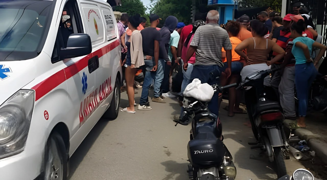 White emergency van with red stripe (ambulance) parked on a street as a crowd gathers nearby, with motorcycles in the foreground and people standing around.
