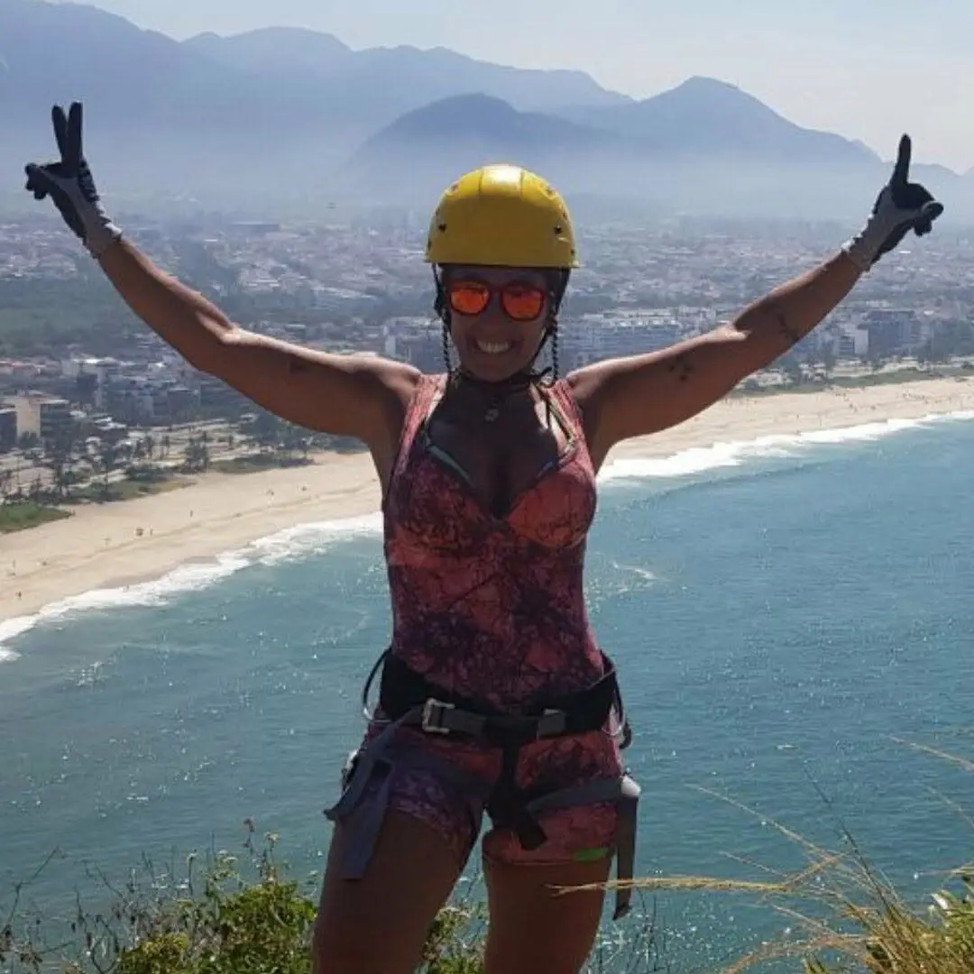 Woman climber on a cliff edge, wearing a yellow helmet, harness, and orange sunglasses, arms raised in victory with the ocean beneath.