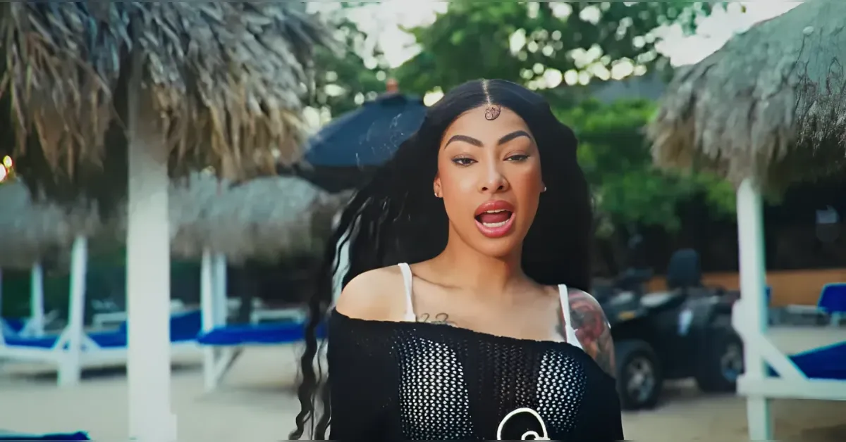 Woman with long dark hair wearing an off-shoulder black mesh top, speaking or singing on a sunny tropical beach with thatched umbrellas in the background.
