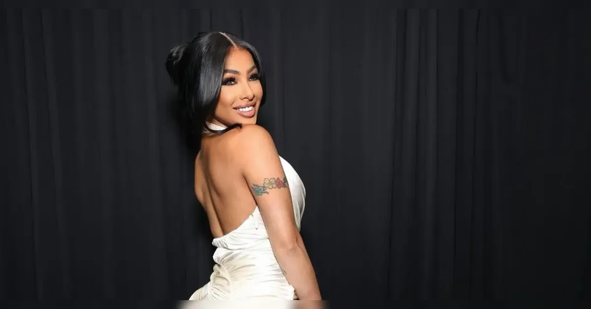 Woman in a white backless gown turns with a smile, against a dark curtain backdrop; tattoo on her upper arm visible from the side view.