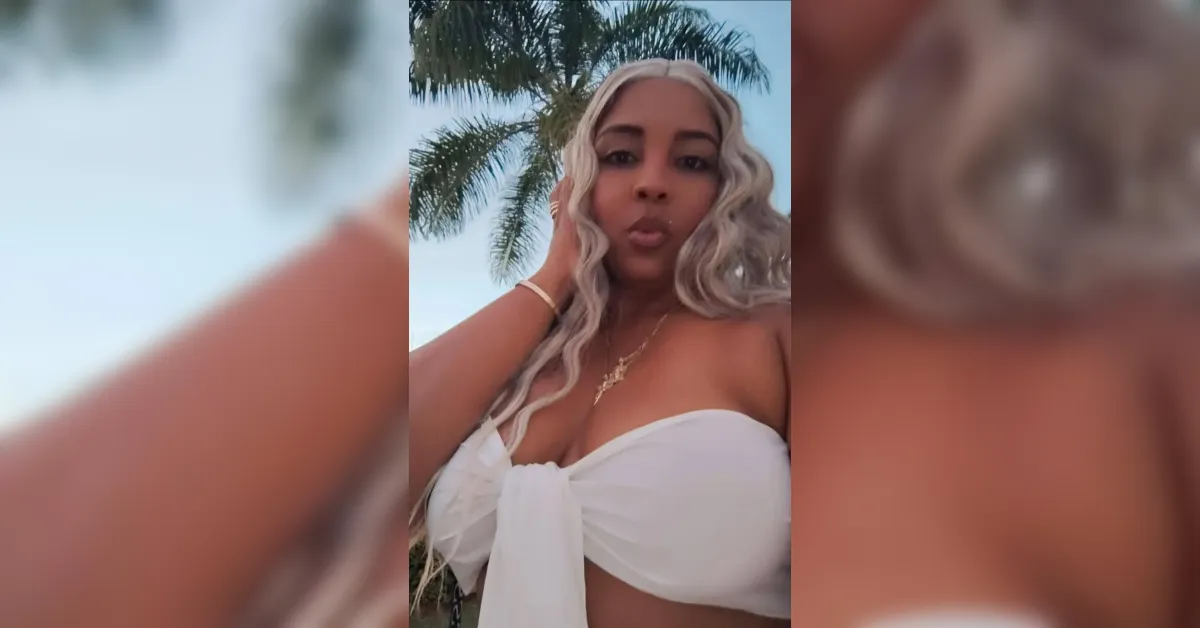 Selfie video frame of a woman with long blonde curls wearing a white strapless top, palm trees in the background.
