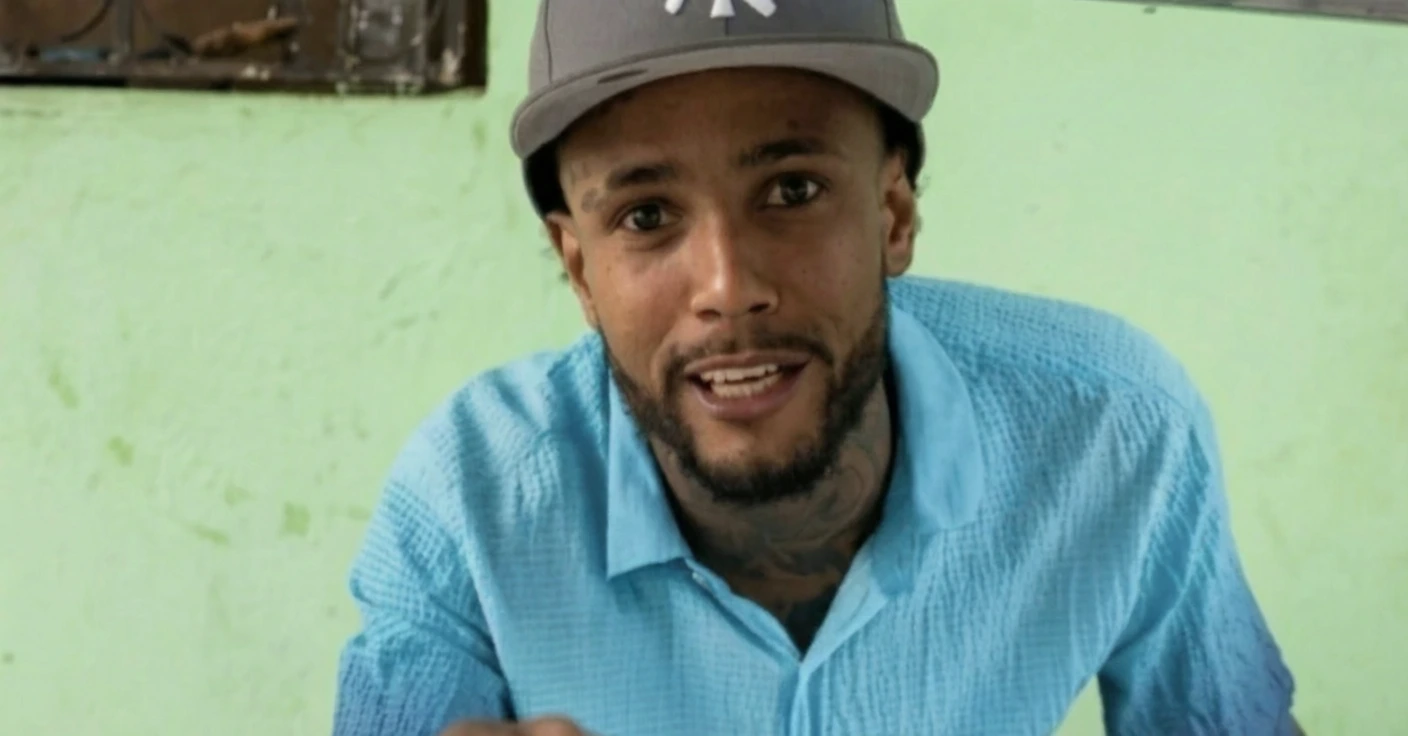 Portrait of a man wearing a gray cap and a blue textured shirt, smiling at the camera against a light green wall.