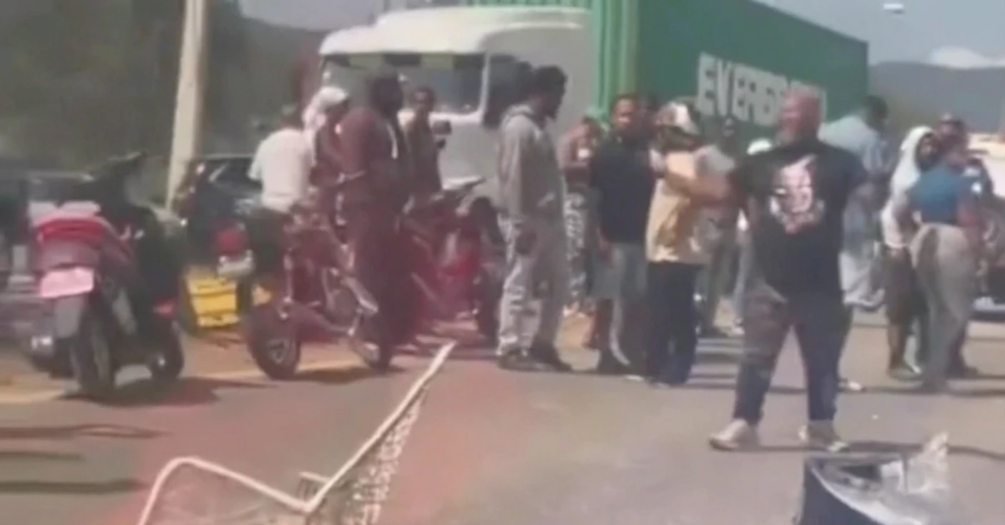 Crowd of bystanders on a sunlit street near a white truck and a green trailer, with several motorcycles nearby.