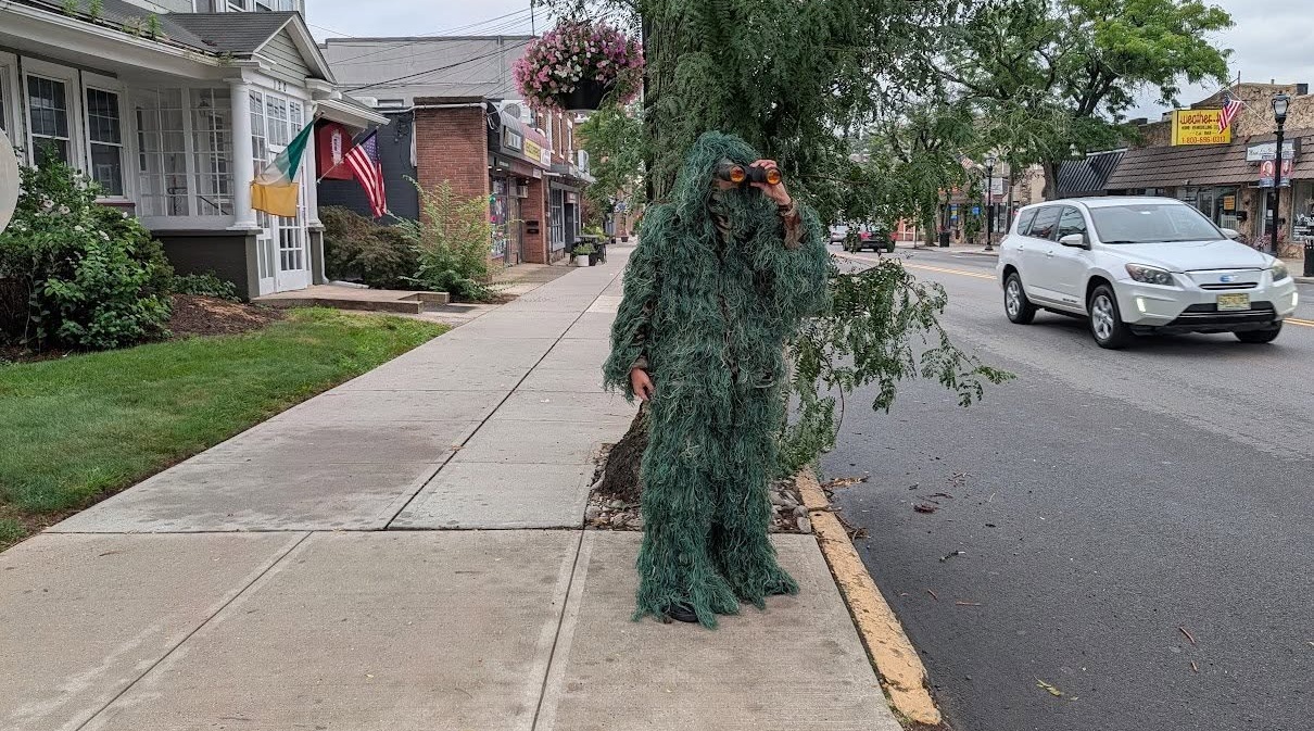 Person in a green ghillie suit stands on a sidewalk in a small town, looking through orange binoculars.