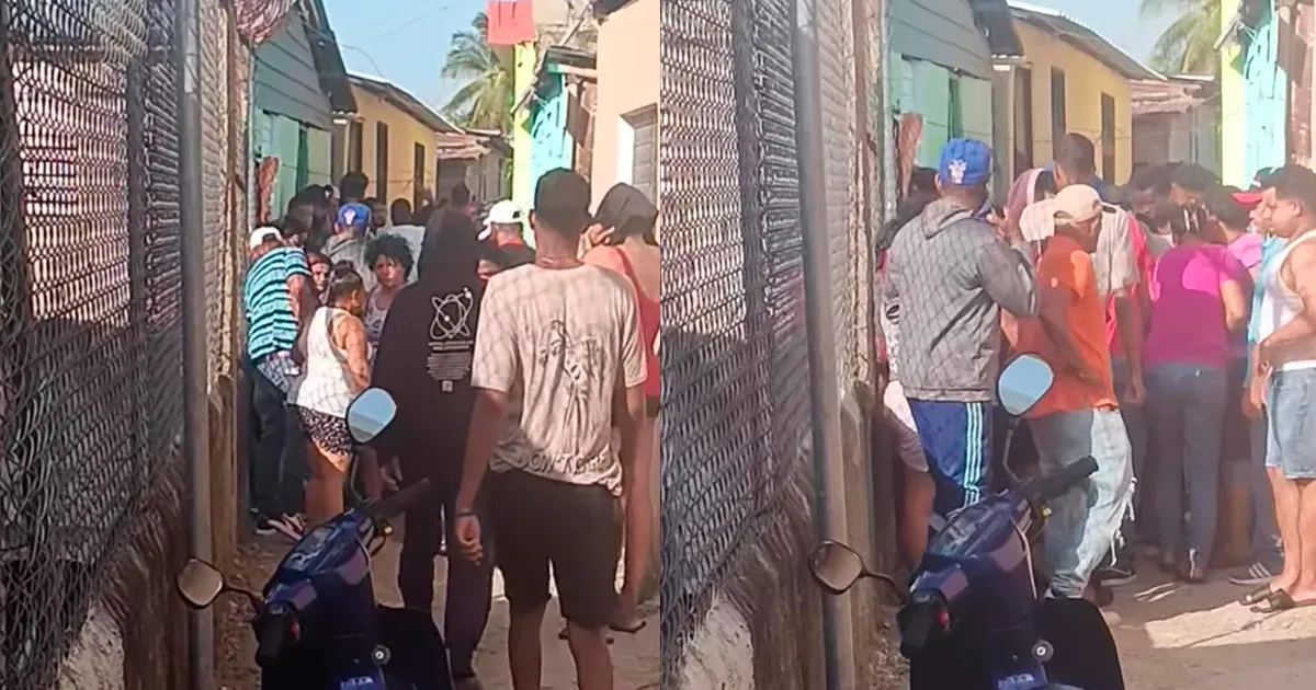 Crowded narrow alleyway with a chain-link fence on both sides, people standing and talking, a blue scooter in the foreground.