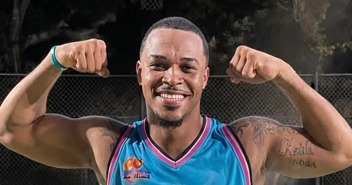 Smiling man wearing teal basketball jersey, flexing his biceps outdoors at night with a chain-link fence behind him.