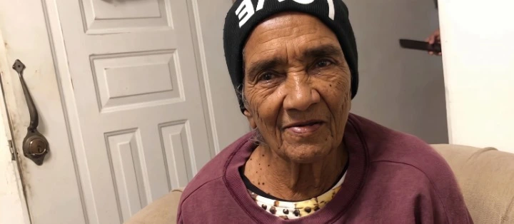 Elderly woman wearing a black beanie and maroon sweatshirt sits on a beige couch, smiling softly at the camera.
