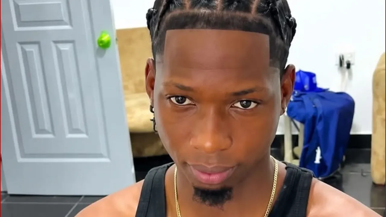 Close-up of a young man with braided hair and a goatee looking directly at the camera indoors, wearing a black tank top and gold chain with a gray door in the background.