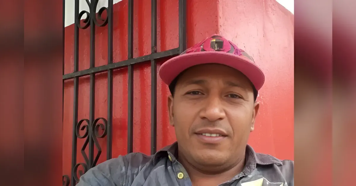 Man wearing a pink cap and gray shirt taking a selfie in front of a red wall with a black metal gate.