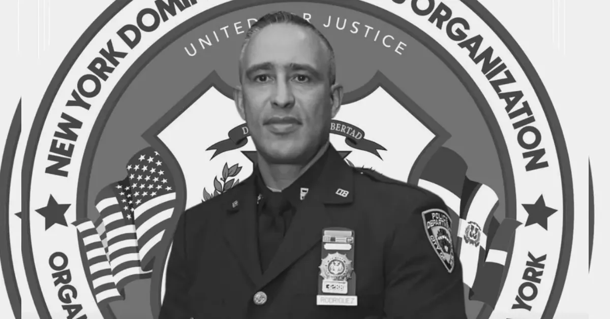 Male police officer in a dress uniform standing in front of a large circular emblem that reads 'New York Dominican Organization' with flags in the background, conveying a formal ceremony setting.