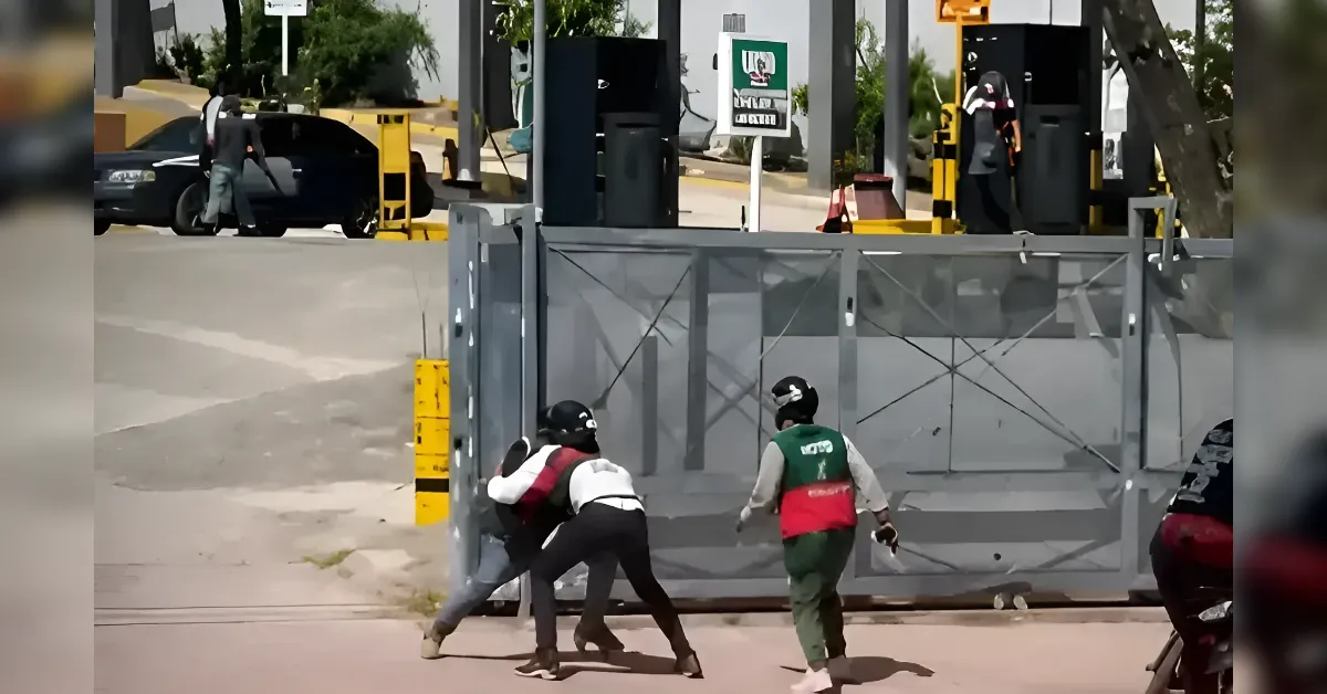 Three people wearing helmets push against a large metal gate at a vehicle checkpoint, with cars and a gas pump visible in the background.