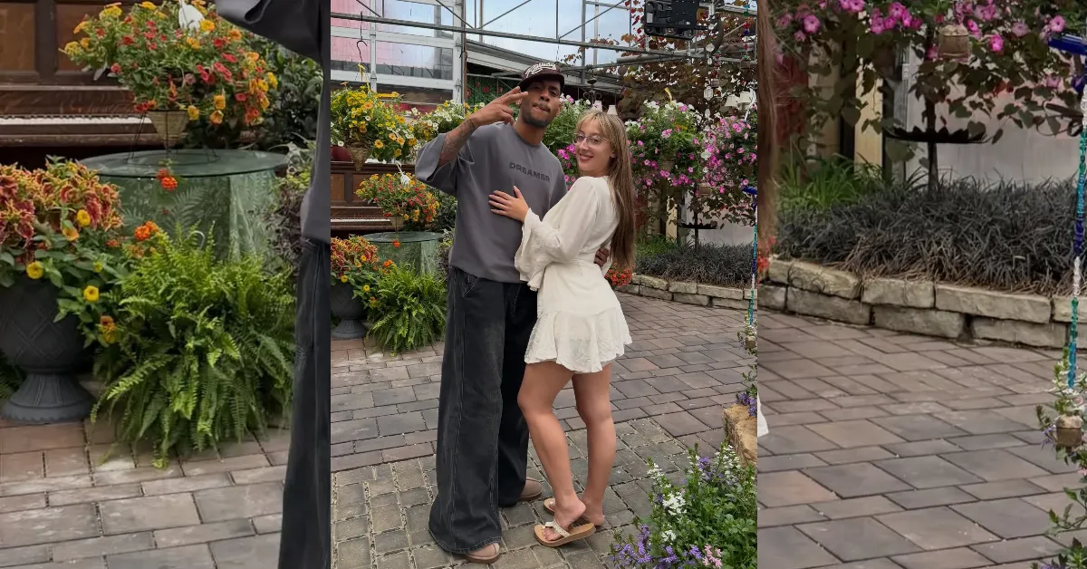 A couple poses together in a flower-filled outdoor garden setting, the man in a gray hoodie and cap, the woman in a white dress, smiling at the camera.