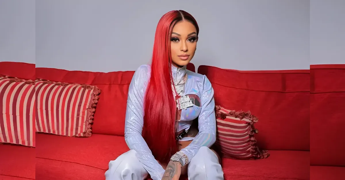 woman with long bright red hair sits on a red sofa in a shiny light-blue outfit with jewelry, facing the camera
