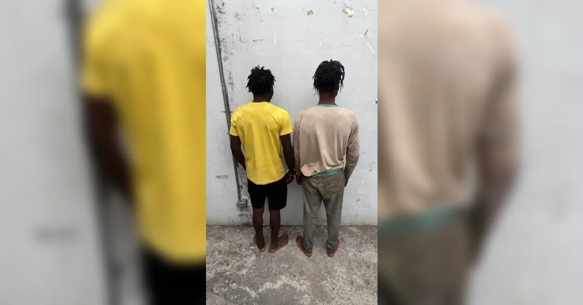 Two men stand with their backs to the camera, hands behind their backs, against a gray wall.