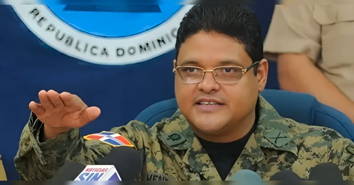 Military officer in camouflage uniform speaking at a press briefing, hand raised as he addresses reporters.