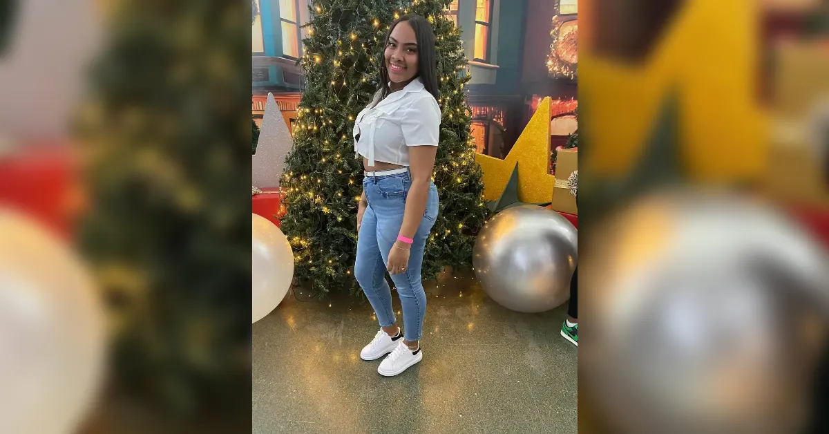 Young woman in a white cropped shirt and jeans posing beside a decorated, lit Christmas tree indoors.
