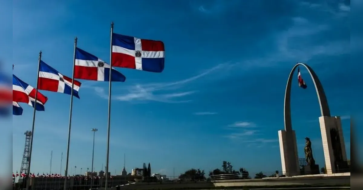 Several flagpoles with red, white, and blue flags fluttering beside a large curved arch monument with a statue at its base, under a blue sky.