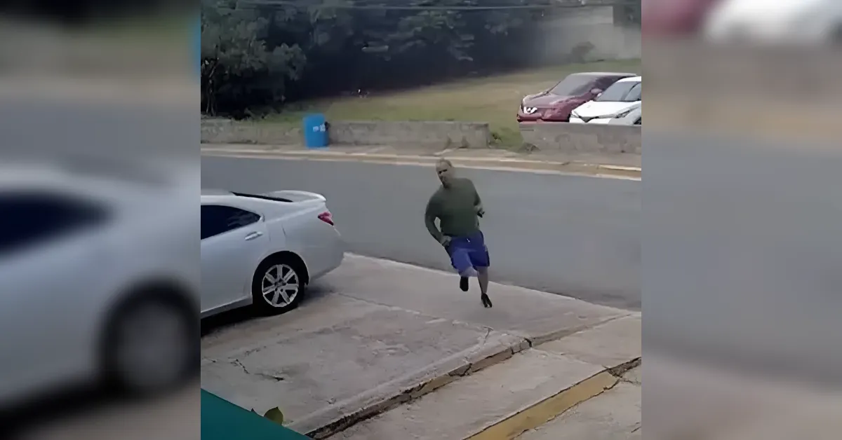 A man in a green shirt and blue shorts sprints along a sidewalk beside a parked white car, with a street and cars in the background
