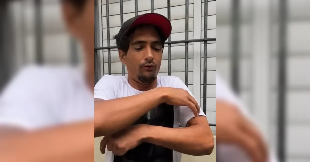 Man wearing a white T-shirt and red cap, looking down while touching his forearm near a metal fence or bars outside a building.