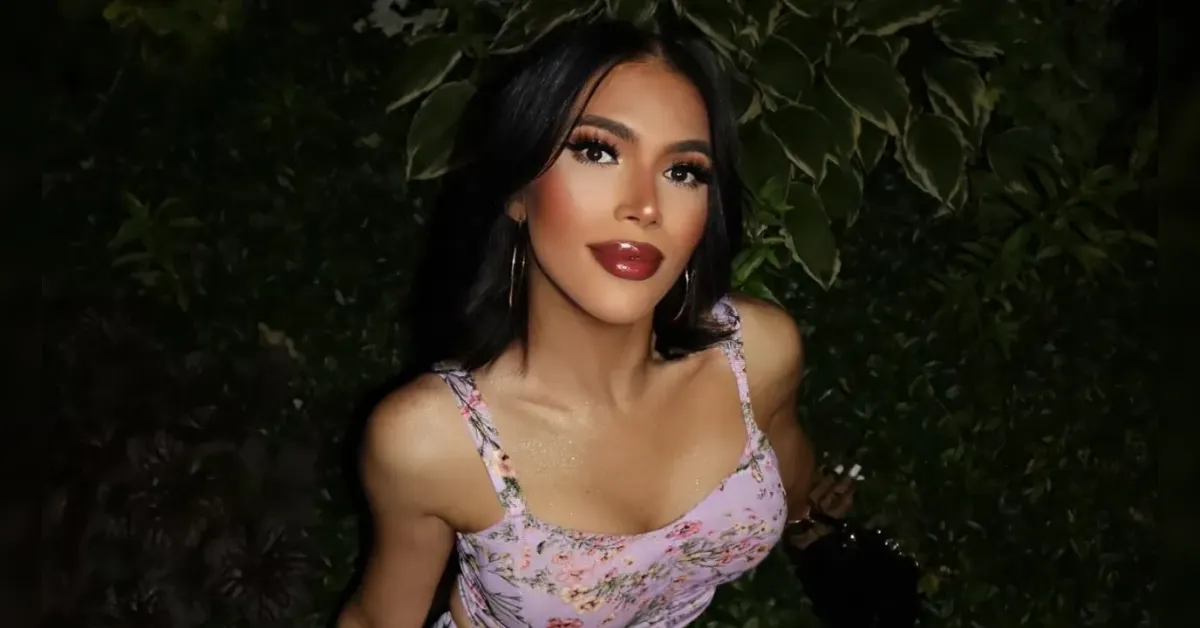 Portrait of a woman with dark hair and bold makeup in a pink floral dress, leaning toward the camera against green foliage at night?conte(req)