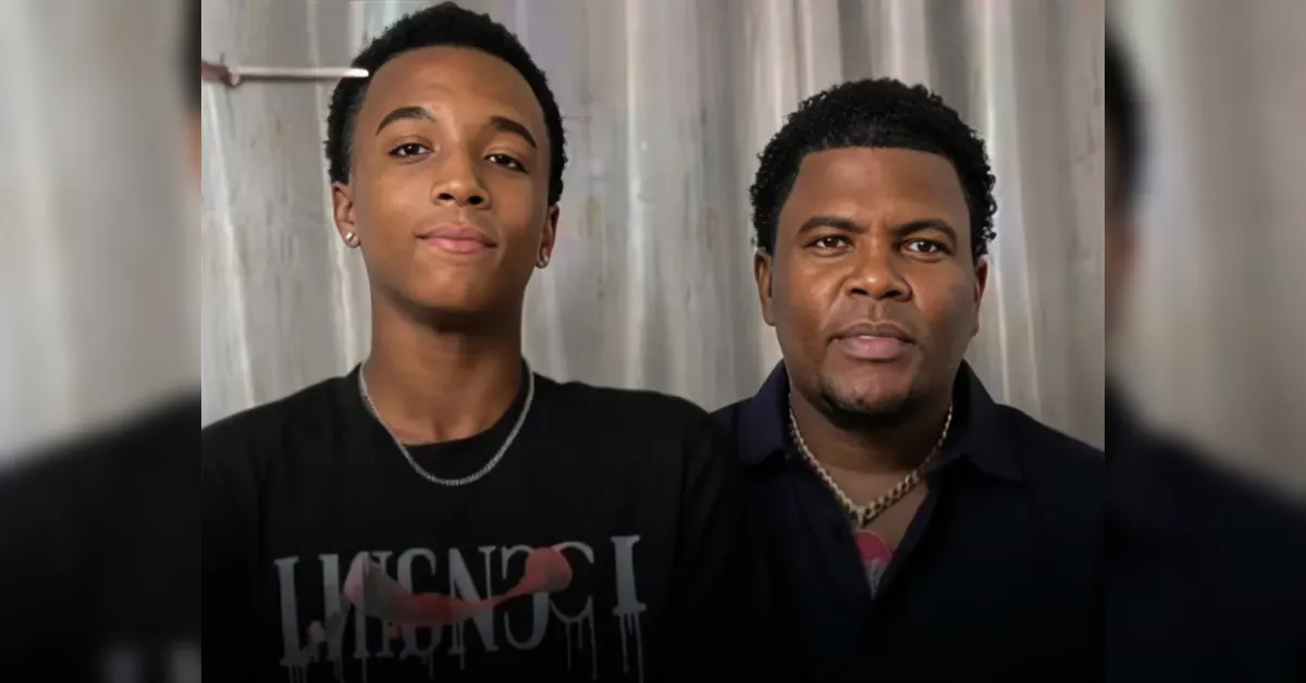 Two men pose for a close-up selfie indoors, standing side by side in front of a light curtain backdrop, one wearing a necklace and a black shirt, both looking at the camera.