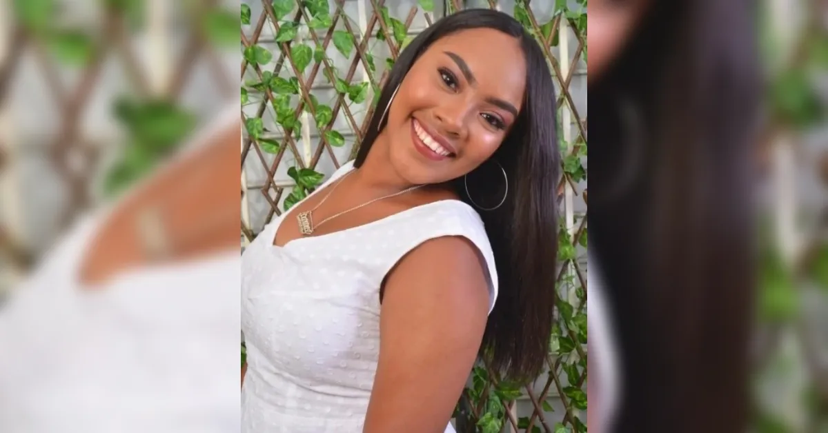 Smiling young woman with long dark hair wearing a white dress, posing outdoors in front of a leafy lattice fence with hoop earrings and a necklace