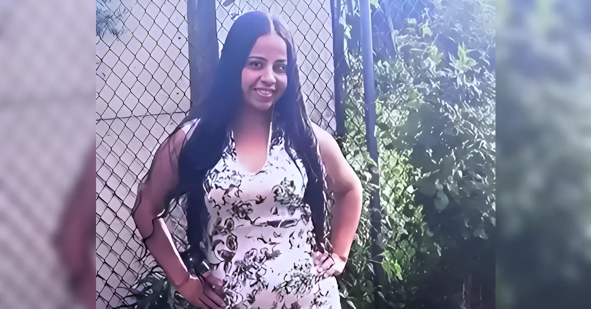 Smiling woman in a white floral dress stands with hands on hips beside a chain-link fence outside placed greenery in the background.