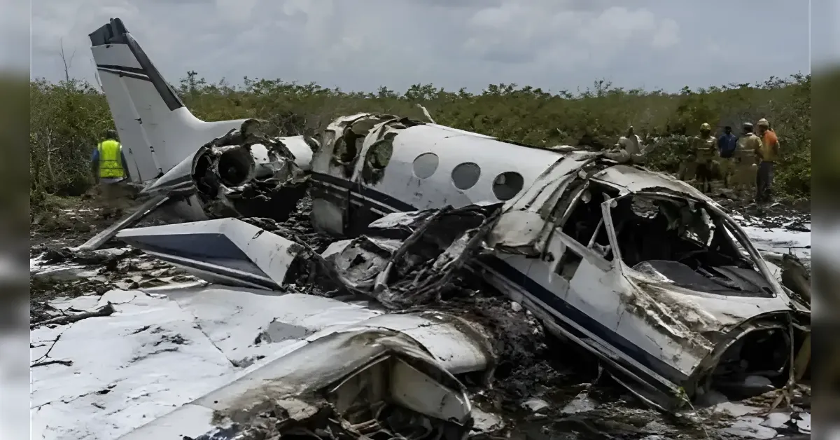 Wrecked white small jet aircraft on a dirt airstrip with twisted fuselage and debris; emergency workers in the background.