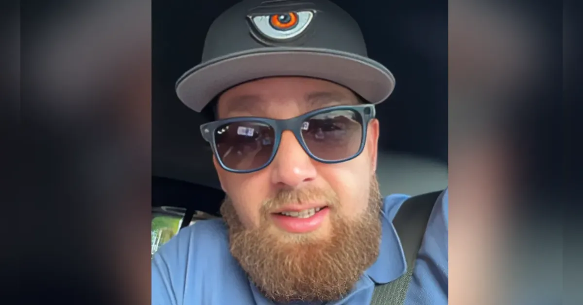 Selfie of a man with a thick beard wearing a gray cap with an orange logo and blue sunglasses inside a car.